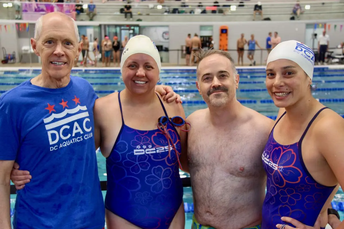 Four DCAC swimmers on a pool deck at a meet, arm in arm and smiling, three in team caps and suits and one in a DCAC T-shirt.