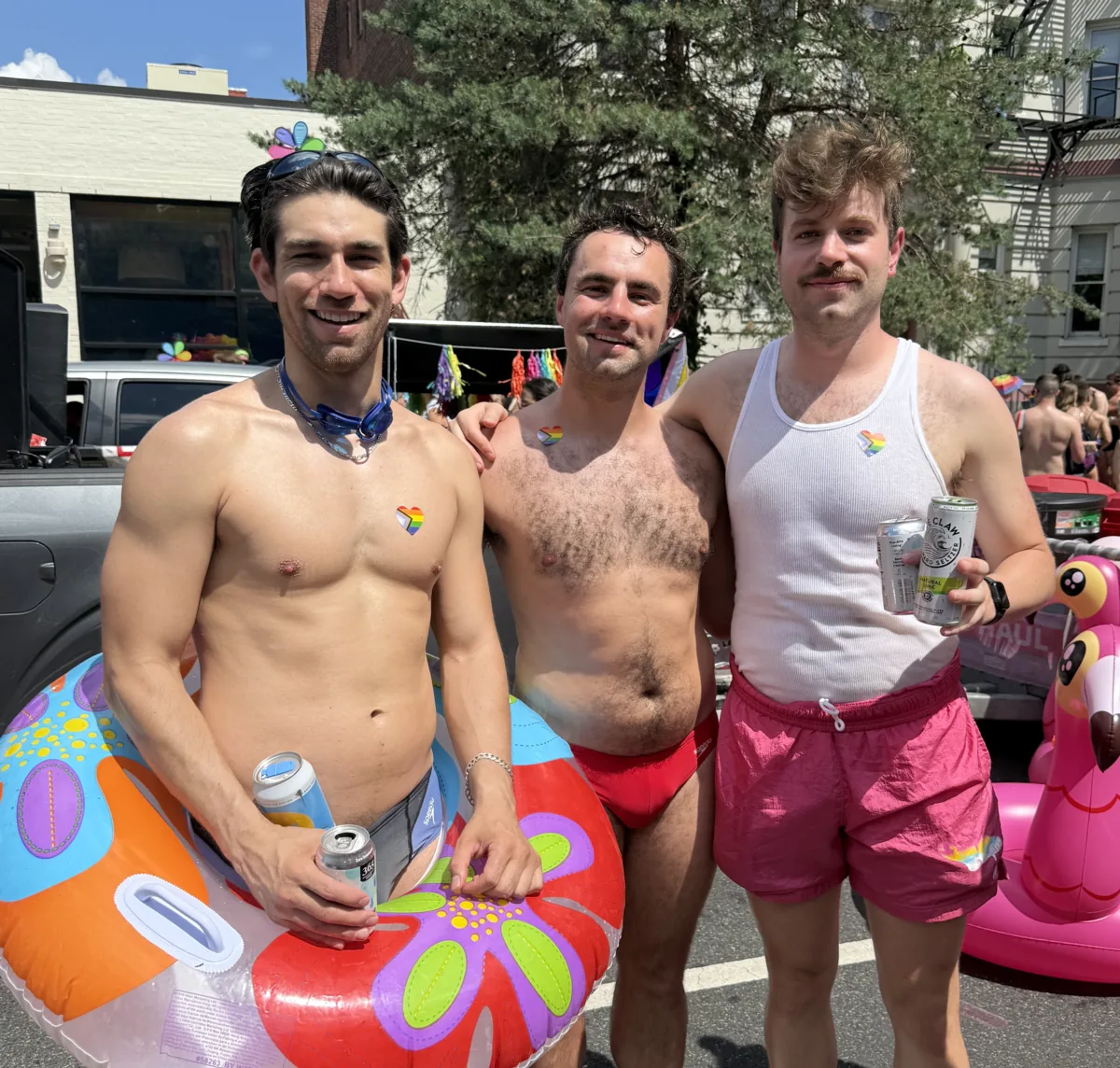 Three DCAC members in swimsuits, holding pool floats and wearing rainbow heart stickers, at a DC Pride event.