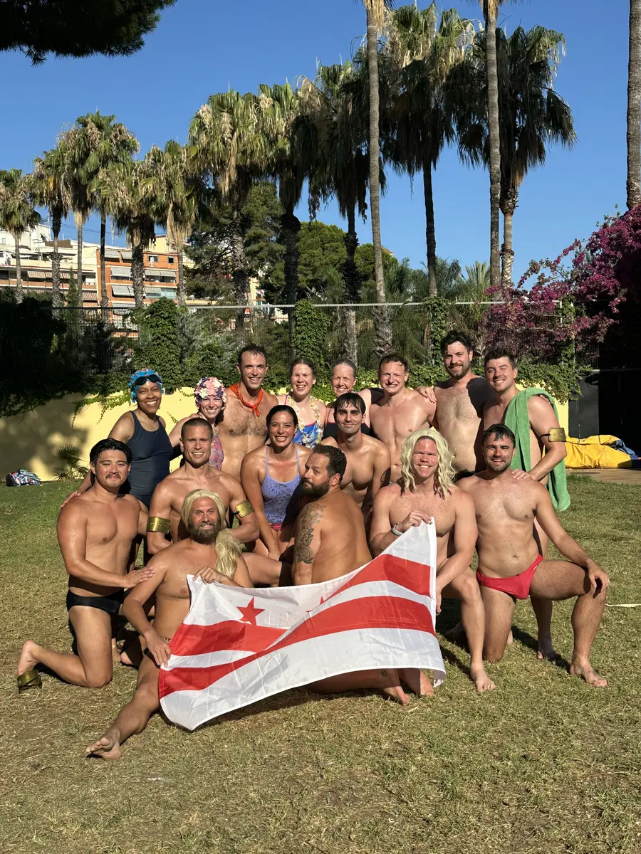 About twenty DCAC swimmers in suits and costume wigs pose on the grass beside an outdoor pool under palm trees, holding up a DC flag.