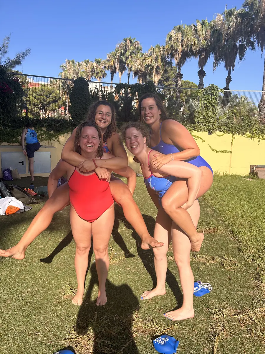 Four smiling DCAC swimmers in pairs, two riding piggyback on the other two, on the grass beside an outdoor pool.